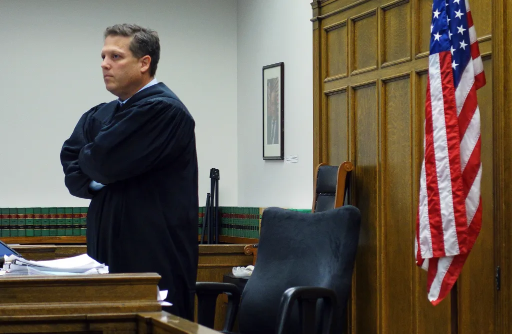 Judge Roger Rogoff in his judicial robe standing in a courtroom, with an American flag to his right.