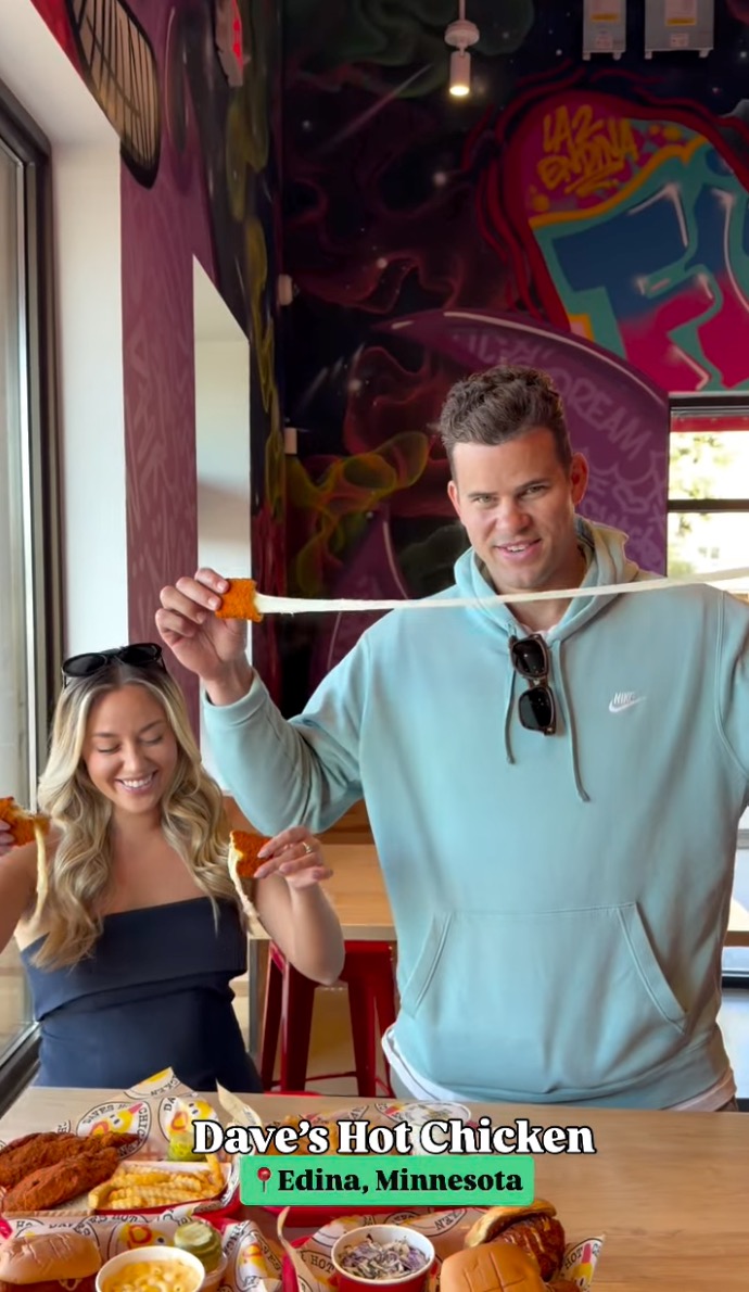 Kris Humphries and a woman holding mozzarella sticks with melted cheese stretching, with Dave's Hot Chicken food on the table.