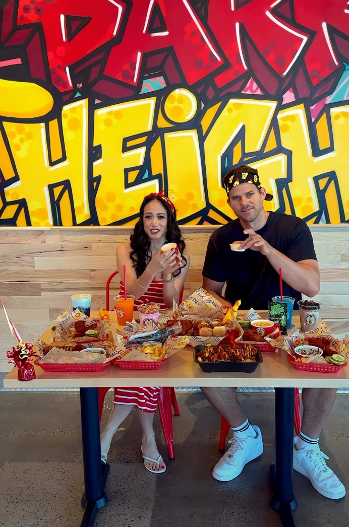 Kris Humphries and a woman sitting at a table with several baskets of chicken and other food items.