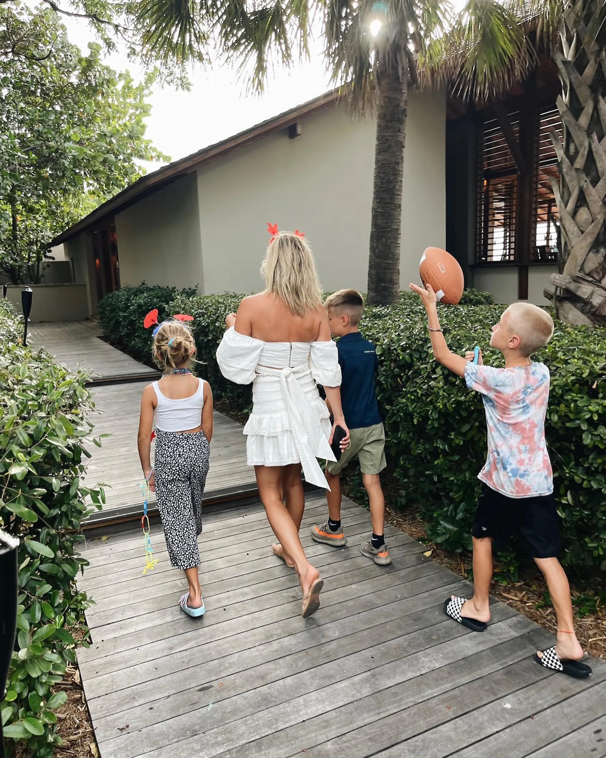 Kristin Cavallari and her children walk on a wooden path surrounded by lush greenery and palm trees, with one son holding a football.