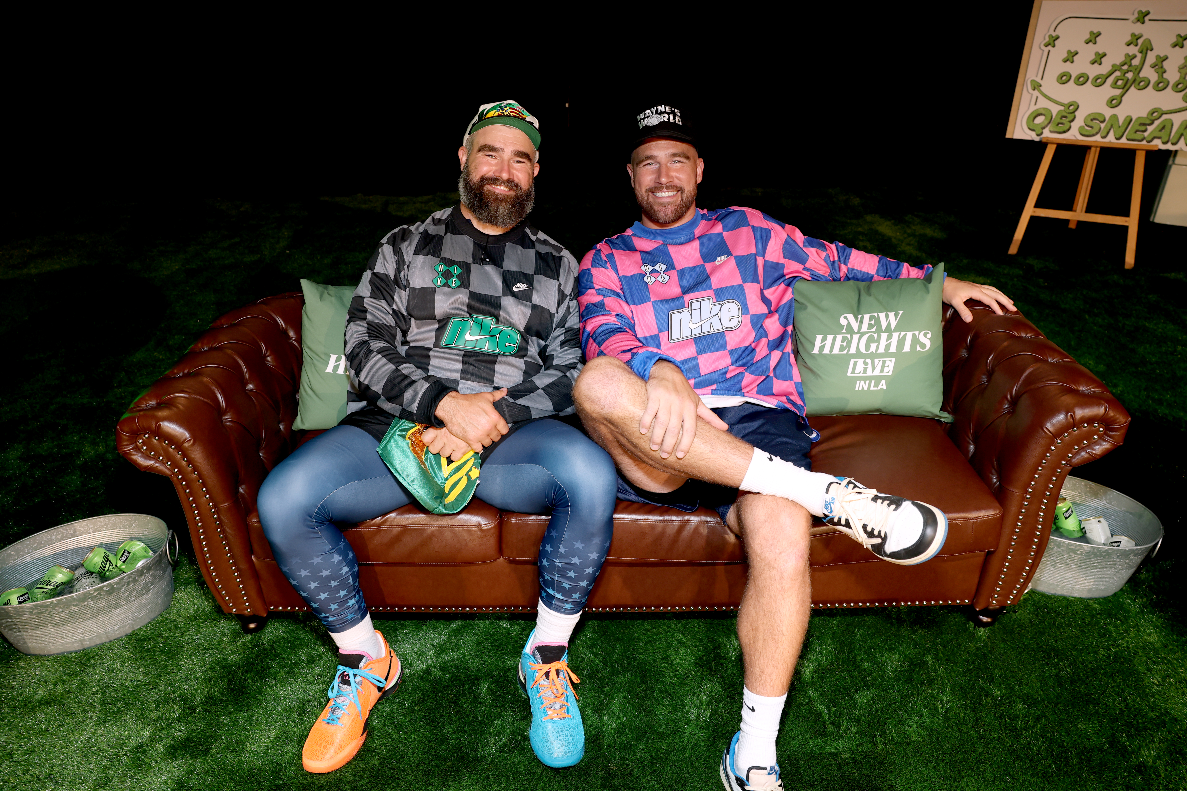 Jason and Travis Kelce smile while sitting on a brown couch at New Heights Live in LA.