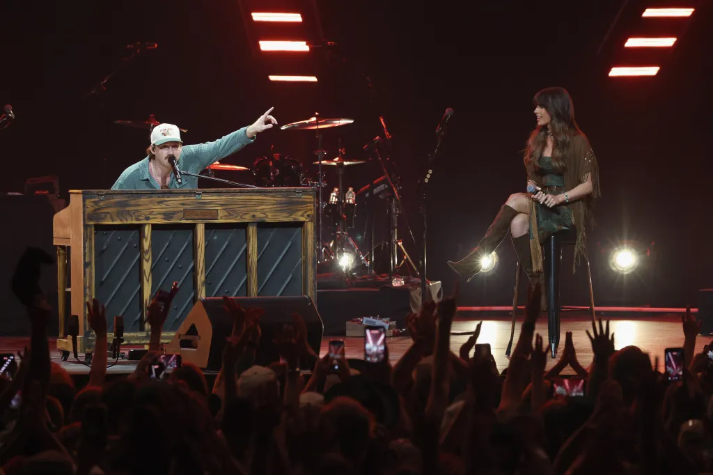 Morgan Wallen at a piano points to Ella Langley sitting on a stool to his right, as they perform for an audience.