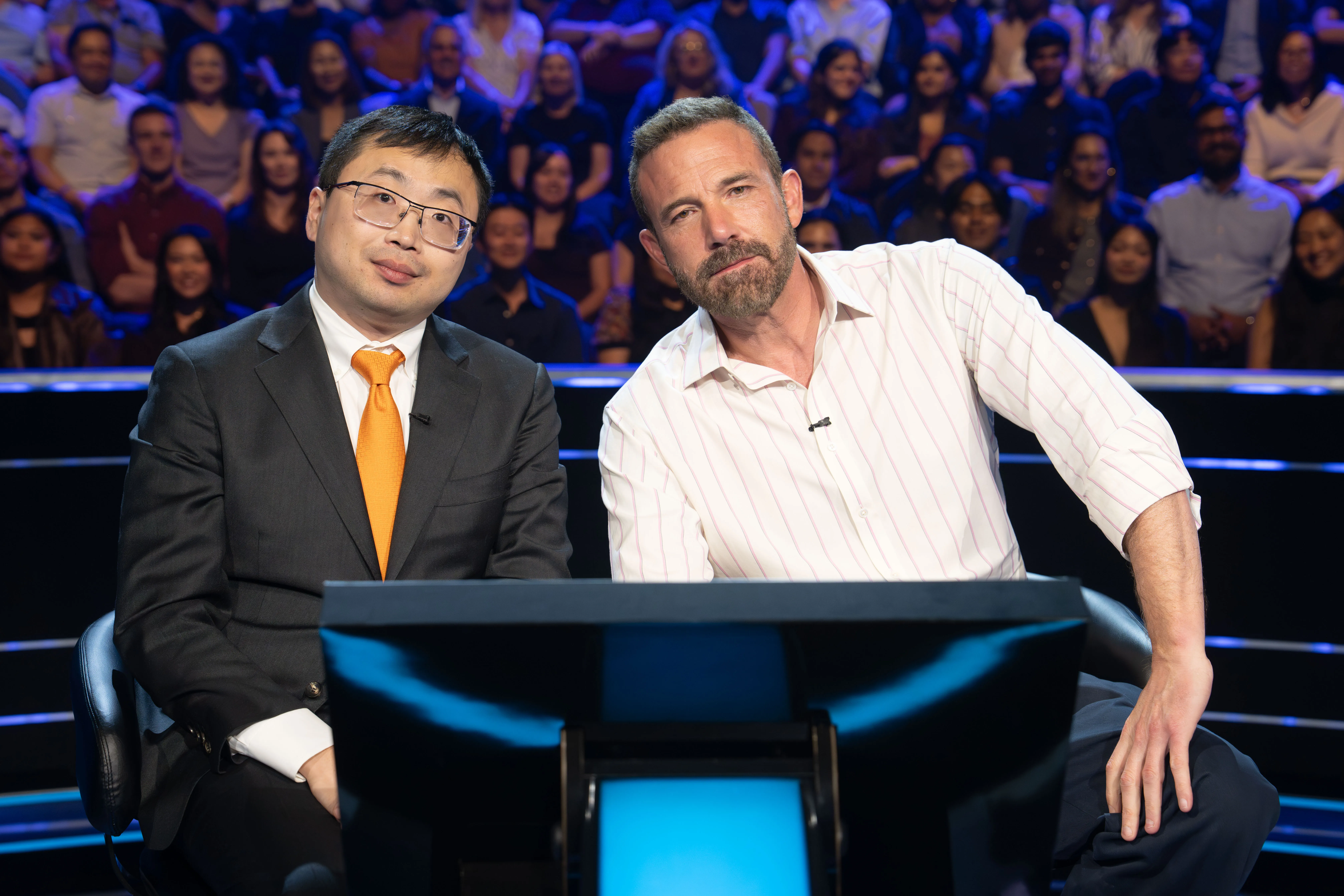 Jamie Ding and Ben Affleck sitting at the "Who Wants to Be a Millionaire?" game show set.