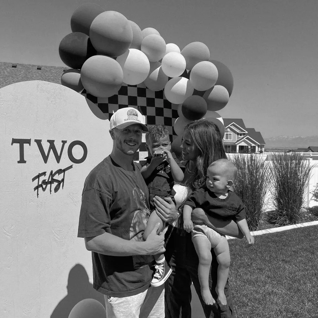 Black and white photo of Layla Taylor and Clayton Wessel holding their two children in front of a "TWO FAST" sign with balloons.