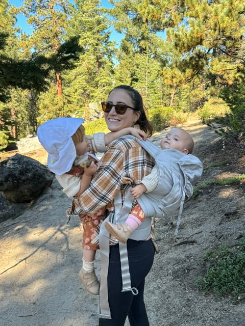 A woman carrying two babies in front and back carriers, smiling at the camera on a wooded trail
