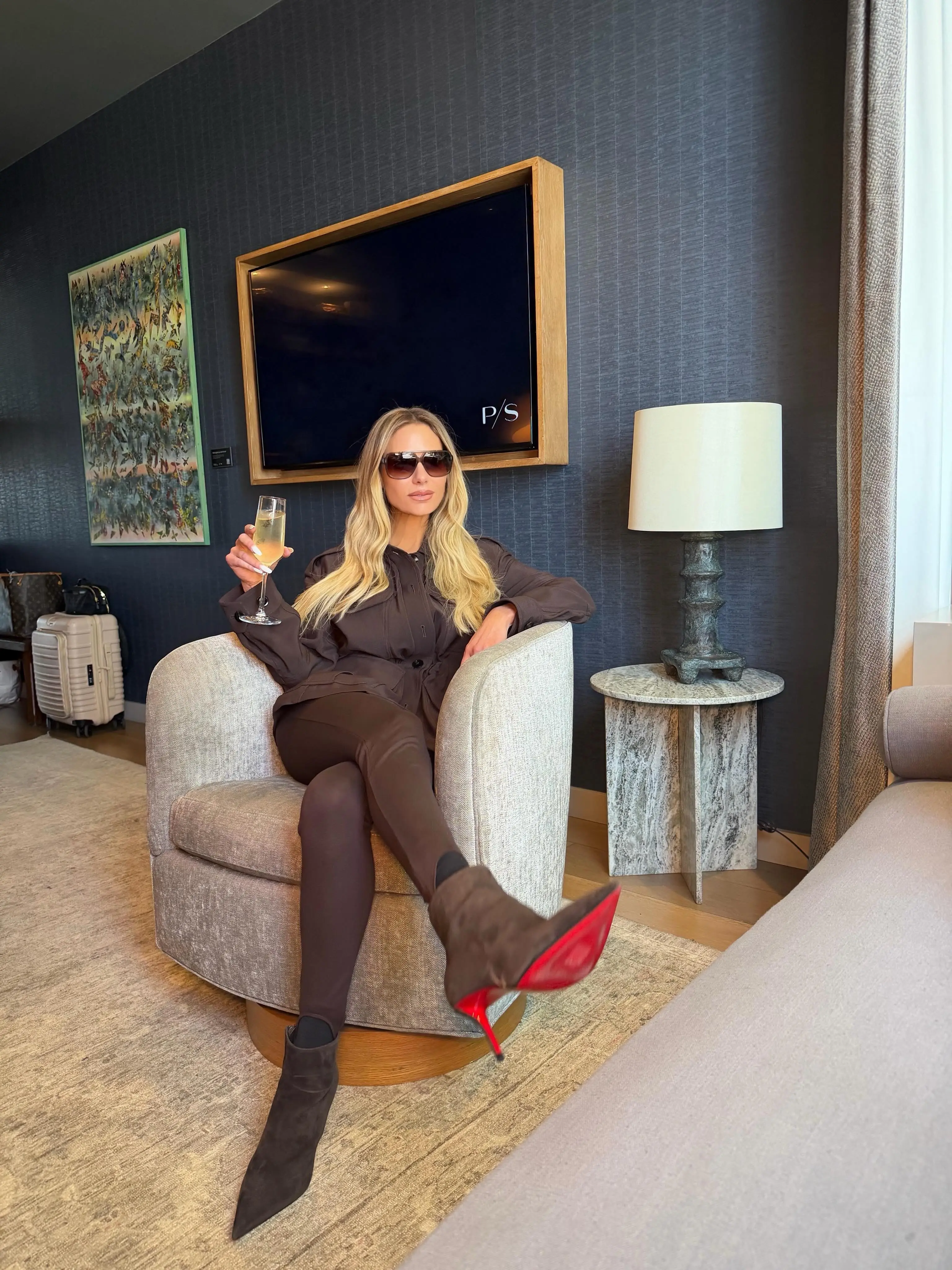 Dorit Kemsley sitting in a chair with champagne, sunglasses, and red-soled high heels, with a suitcase nearby.
