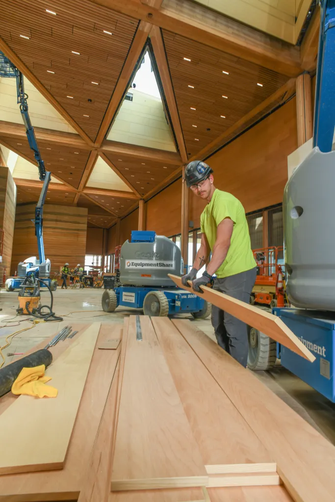 A construction worker in a hardhat and neon yellow shirt carries wood planks in the unfinished Teddy Roosevelt Presidential Library.