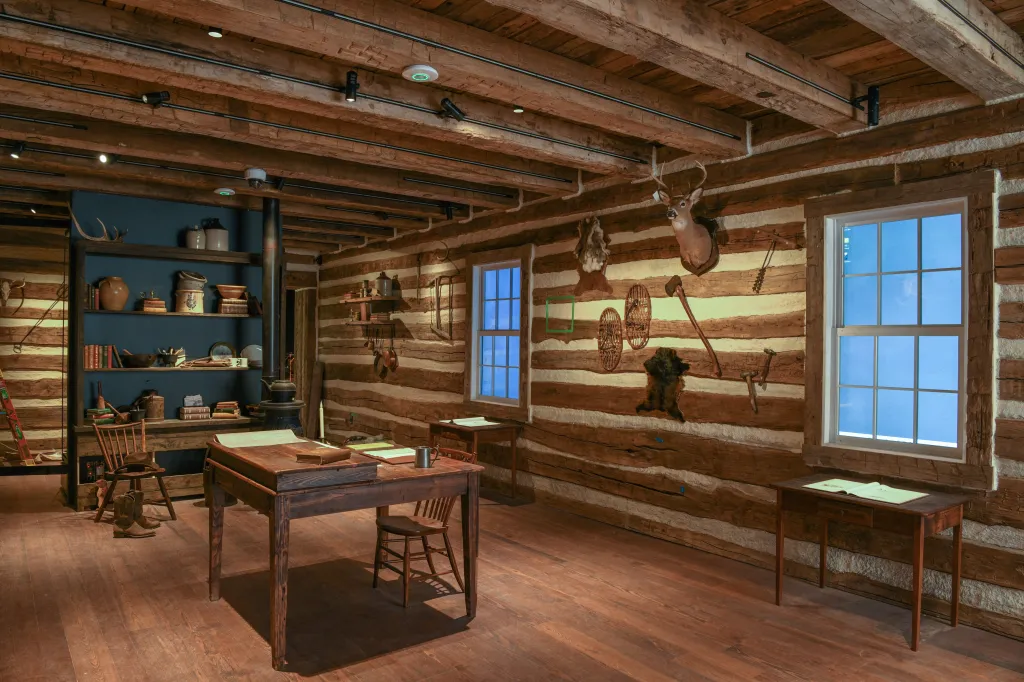 Interior view of a rustic, wood-paneled room with a large wooden desk, shelving with various items, and windows.