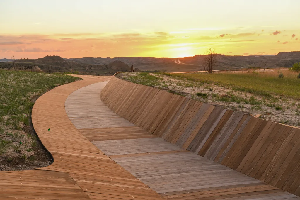 A winding wooden boardwalk in North Dakota at sunset, with hills and plains in the background.