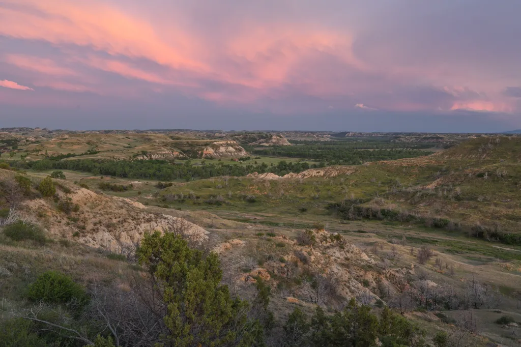 A sweeping landscape of the North Dakota badlands with a green valley, brown hills, and a pink and purple sunset sky.