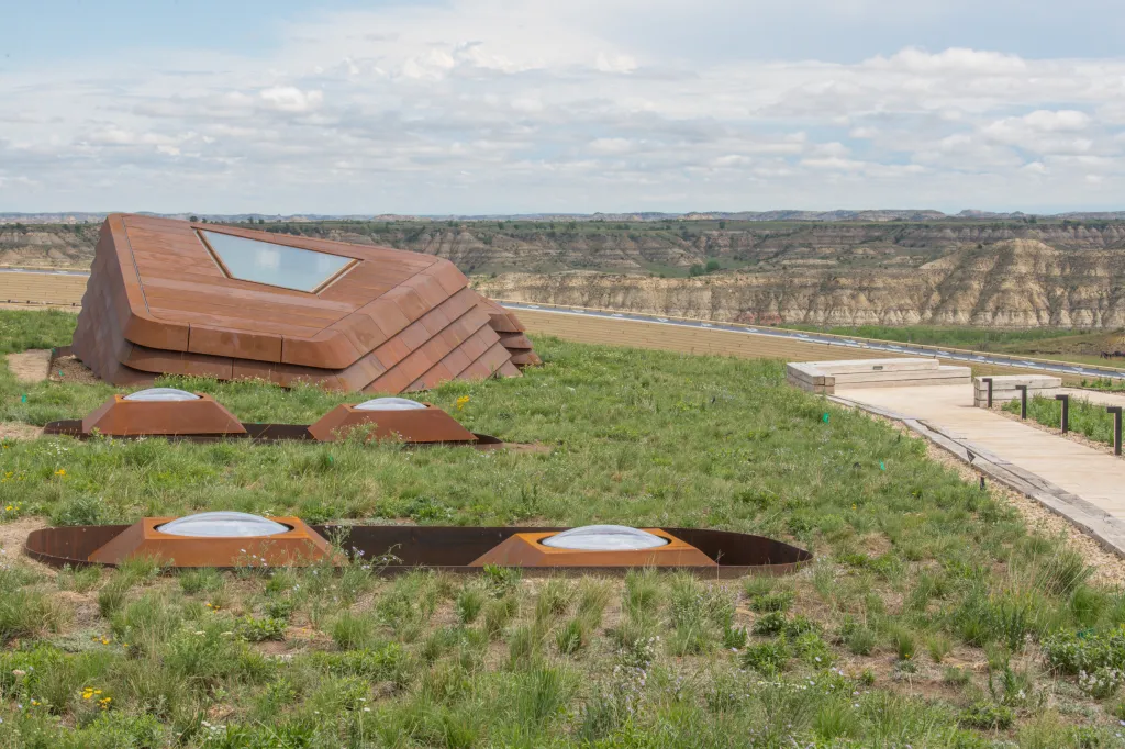 Rooftop of the Teddy Roosevelt Presidential Library in Medora, North Dakota, featuring a large, angular, rusted metal structure with a skylight and several smaller dome-shaped skylights on a grassy roof, overlooking a hilly, arid landscape.