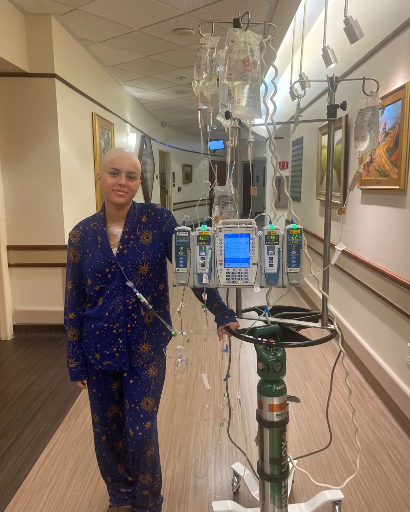 Lilly Dalton, a young woman with a shaved head and wearing blue pajamas, stands in a hospital hallway next to an IV stand.