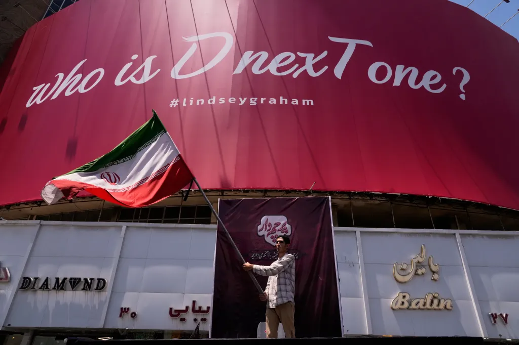 A man waves an Iranian flag in front of a billboard that reads, "Who is D nexT one? #lindseygraham."