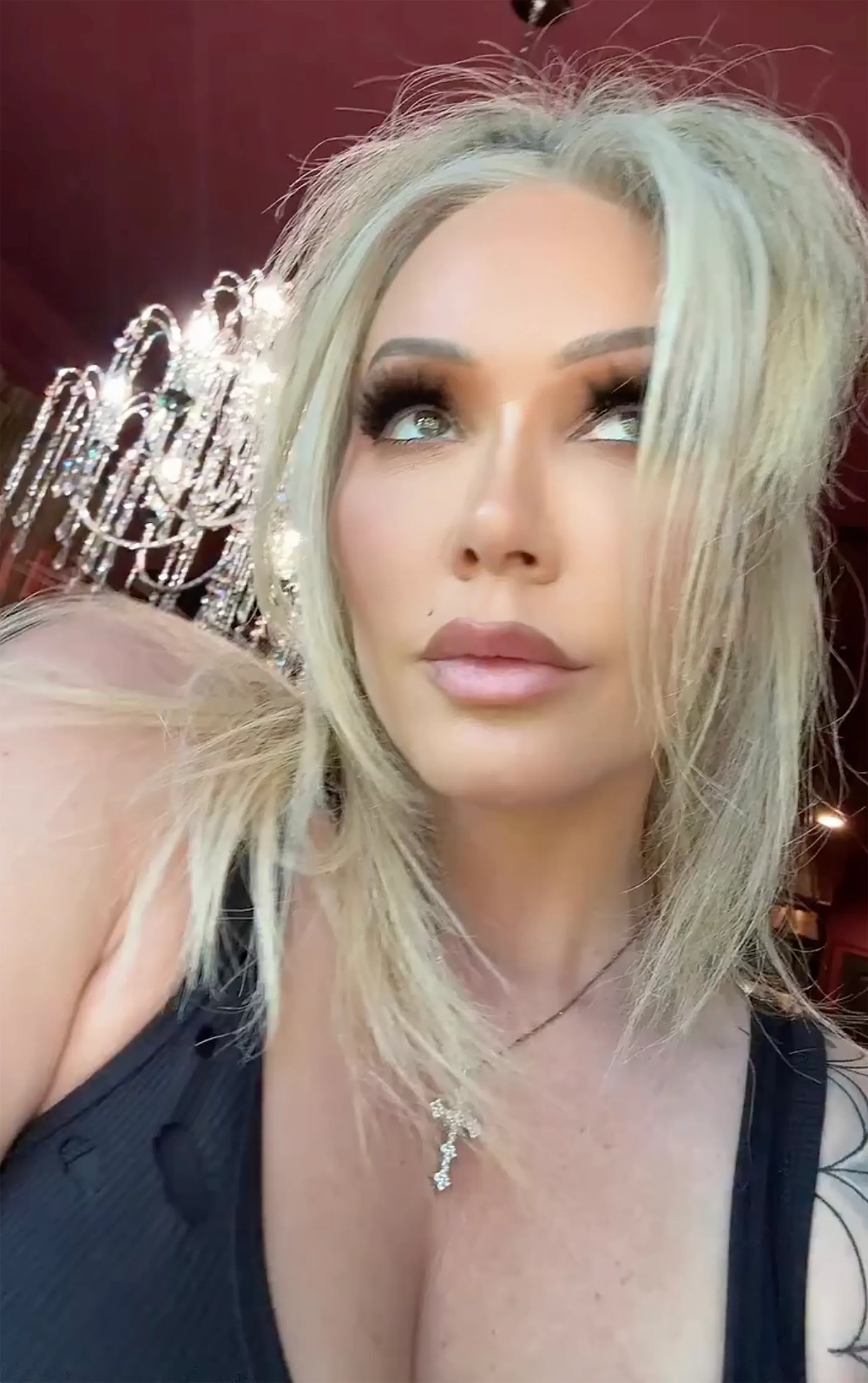 Bunnie XO wearing a black ribbed top and a sparkling cross necklace, looking up and to the right.