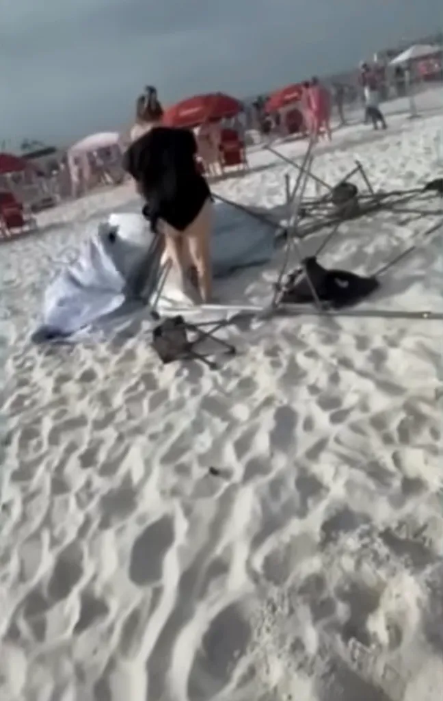 A white beach umbrella has blown over, its metal frame collapsed on the sand, with a person trying to pick it up.