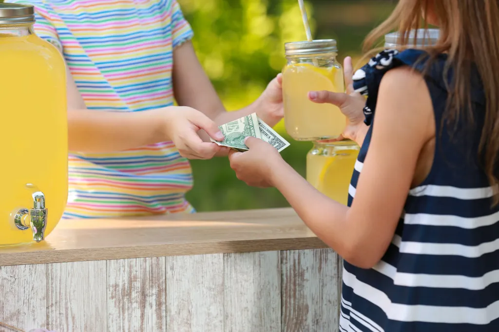 A close-up of a child in a colorful striped shirt taking money from another child in a blue and white striped shirt for a glass of lemonade with a straw.