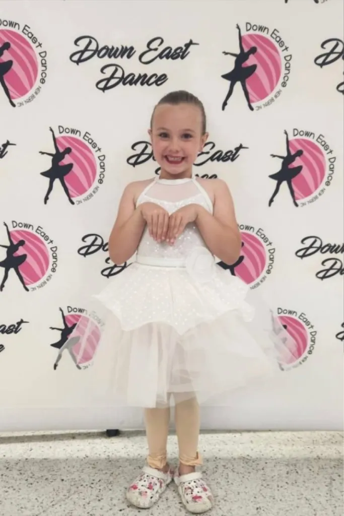 Calli Toler, a 6-year-old girl, smiling in a white dance costume in front of a "Down East Dance" backdrop.