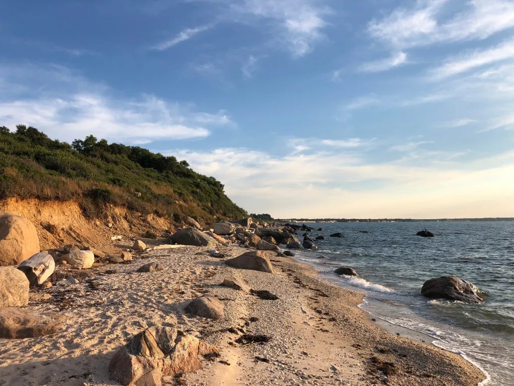 The shore of Inlet Pond County Park in Greenport, Long Island, New York, with a sandy and rocky beach, green hills, and the ocean under a blue sky.
