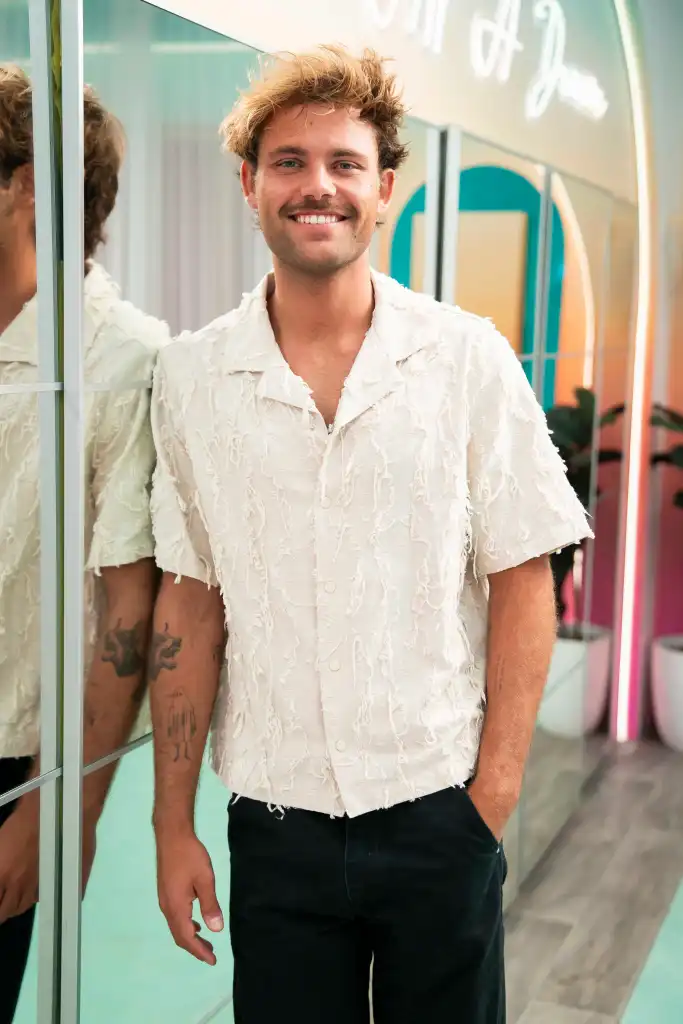 Gal Tshnieder smiling, wearing a white shirt and black pants, with a reflection of himself and his tattoos in a mirror.