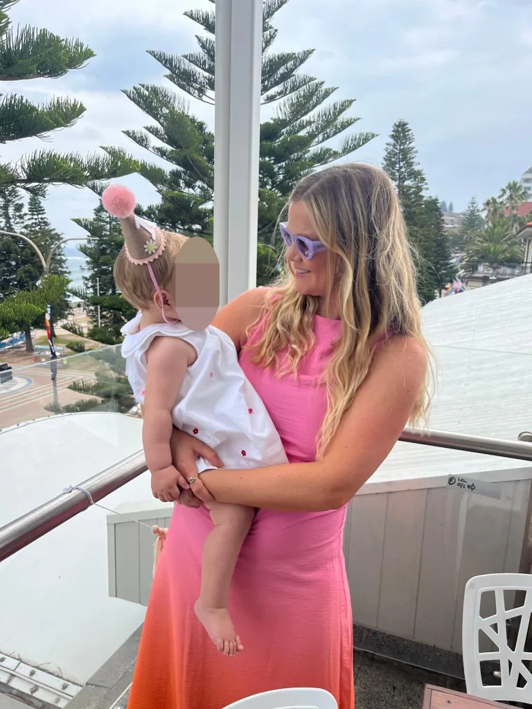 Leah Stewart holding her baby in a birthday hat on a balcony overlooking the ocean.