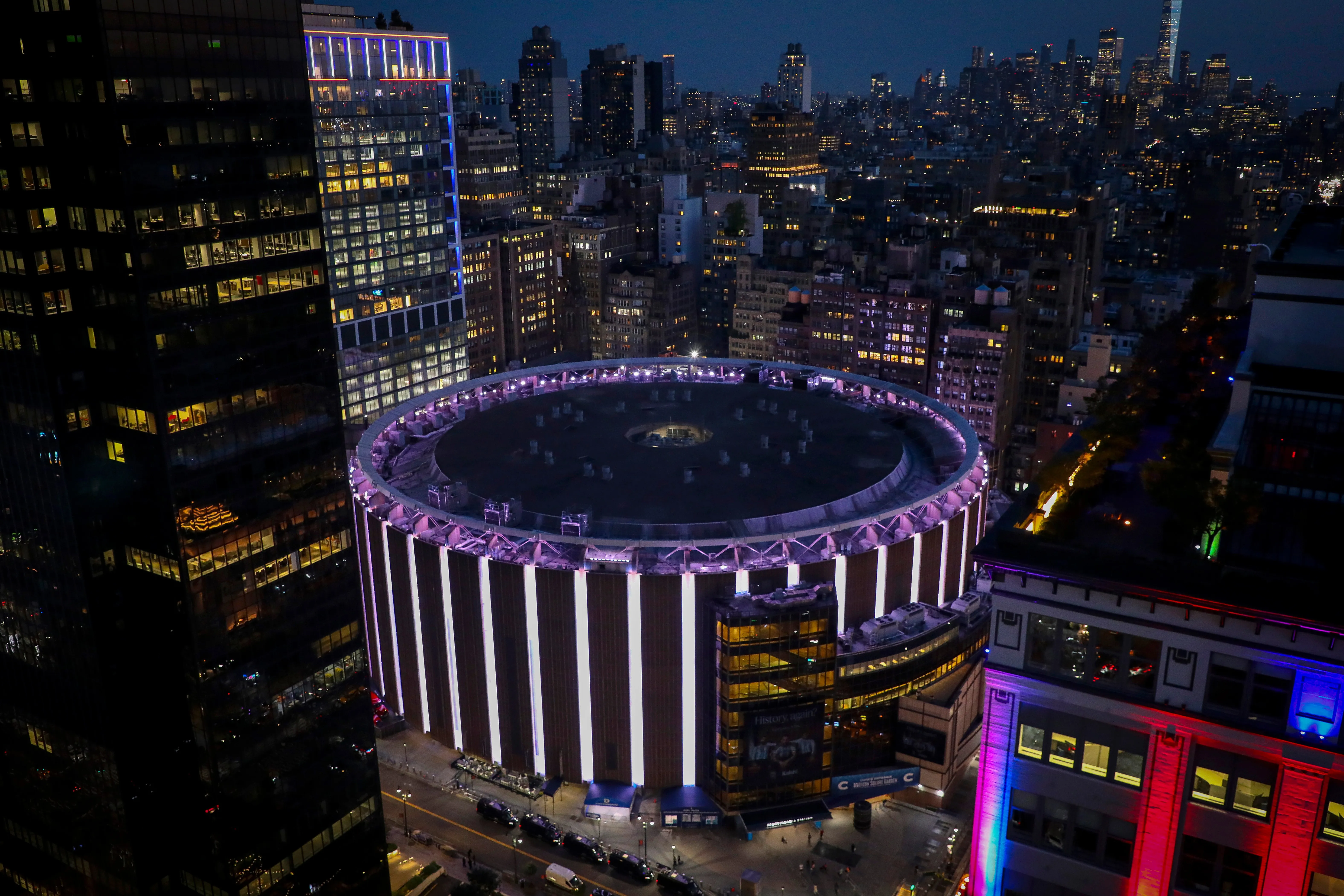 Madison Square Garden at night, illuminated by purple and white lights, surrounded by other New York City buildings.