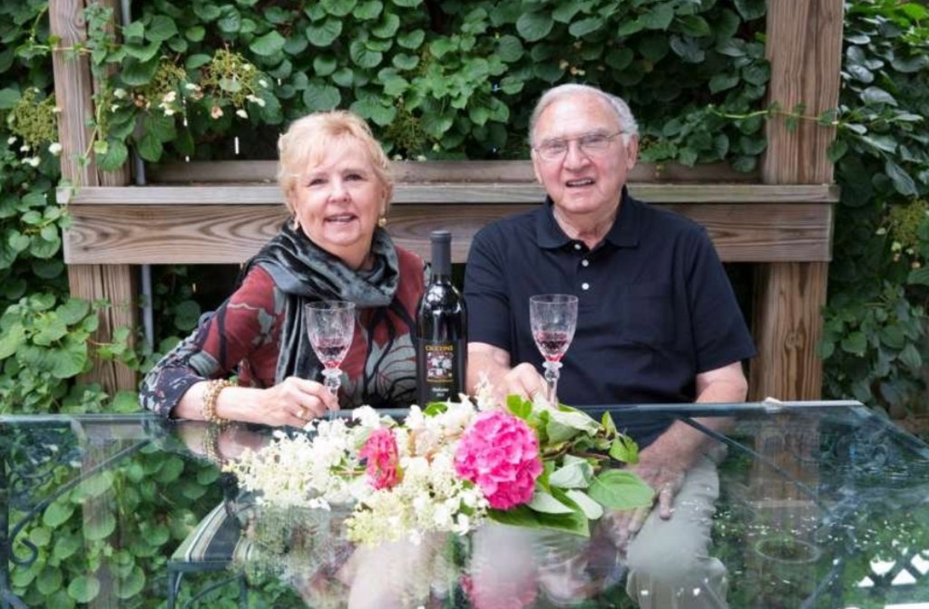 Madonna's stepmother Joan Ciccone and her father Silvio Ciccone sitting at an outdoor glass table with wine and flowers.