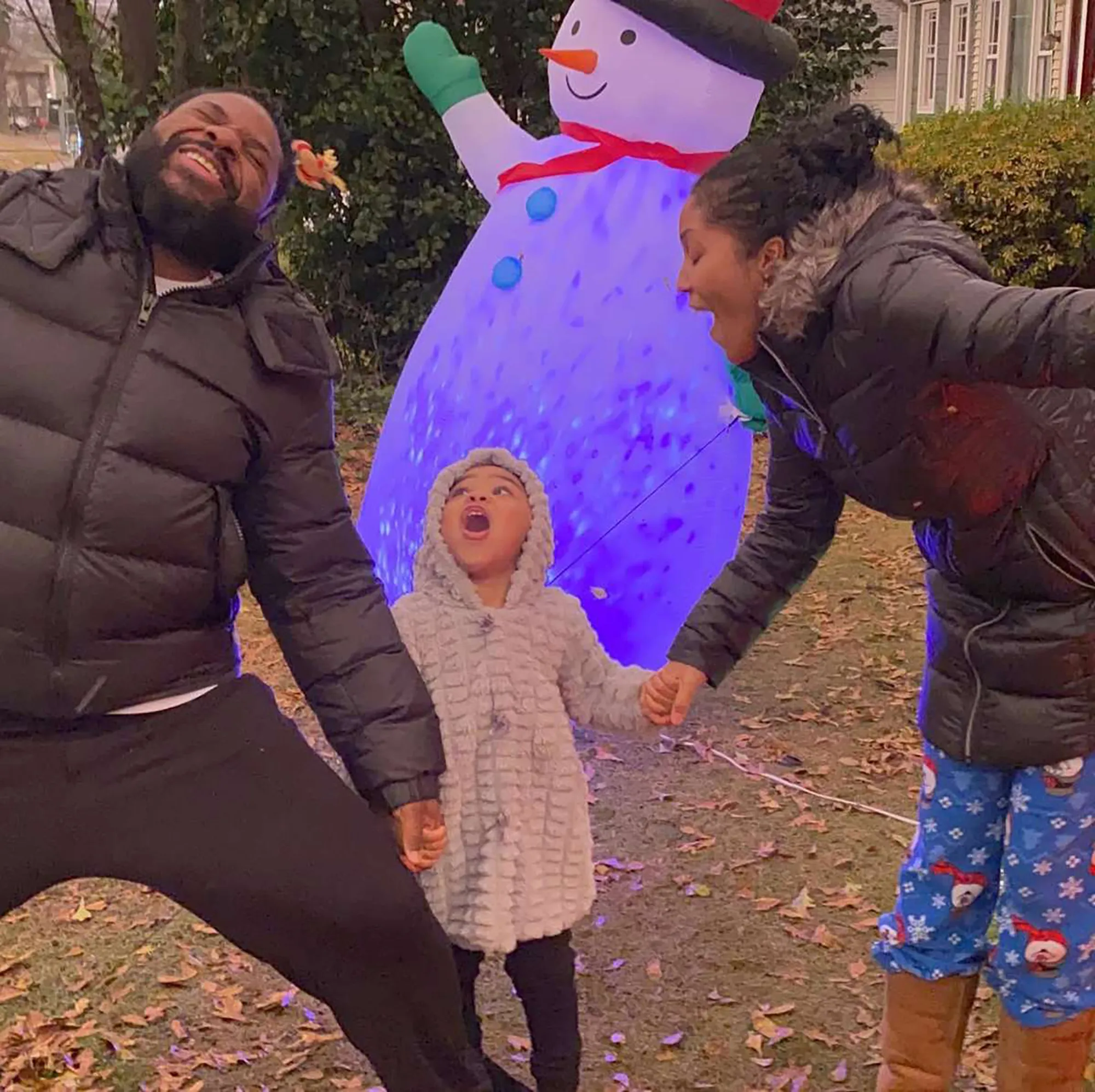 Malcolm-Jamal Warner with his wife and daughter holding hands, laughing next to an inflatable snowman.