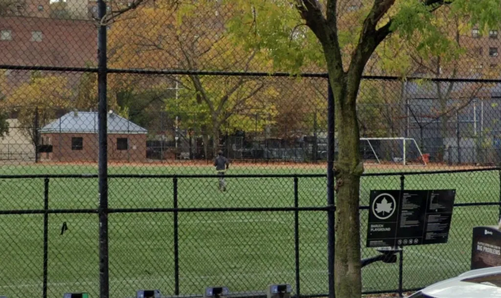 Playground with green turf behind a chain-link fence, with a building, soccer goal, and trees visible.