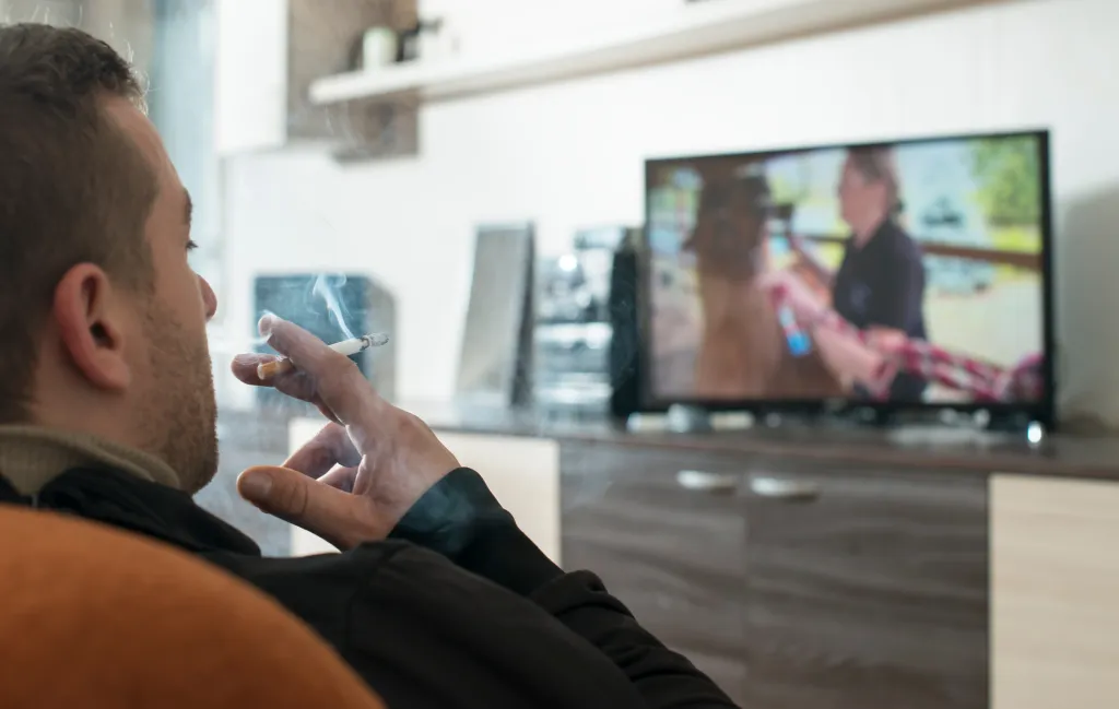Hand of a man smoking a cigarette with the TV on in front of him.