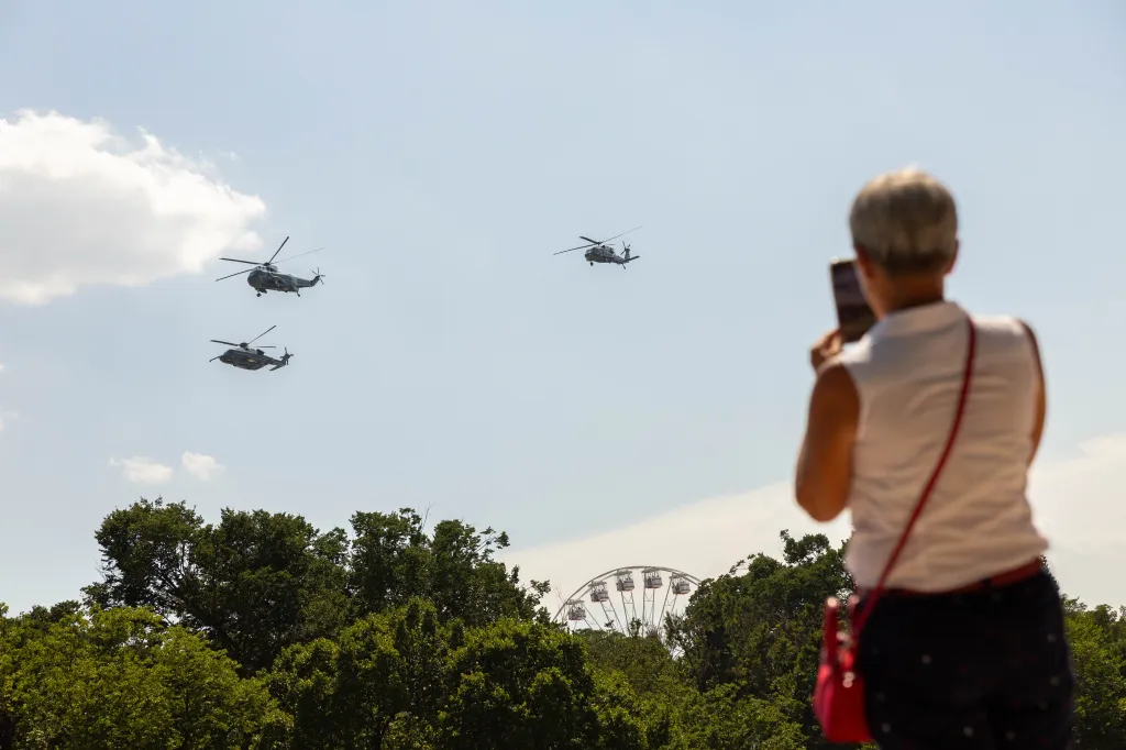 A person photographs military helicopters flying over treetops near a Ferris wheel.