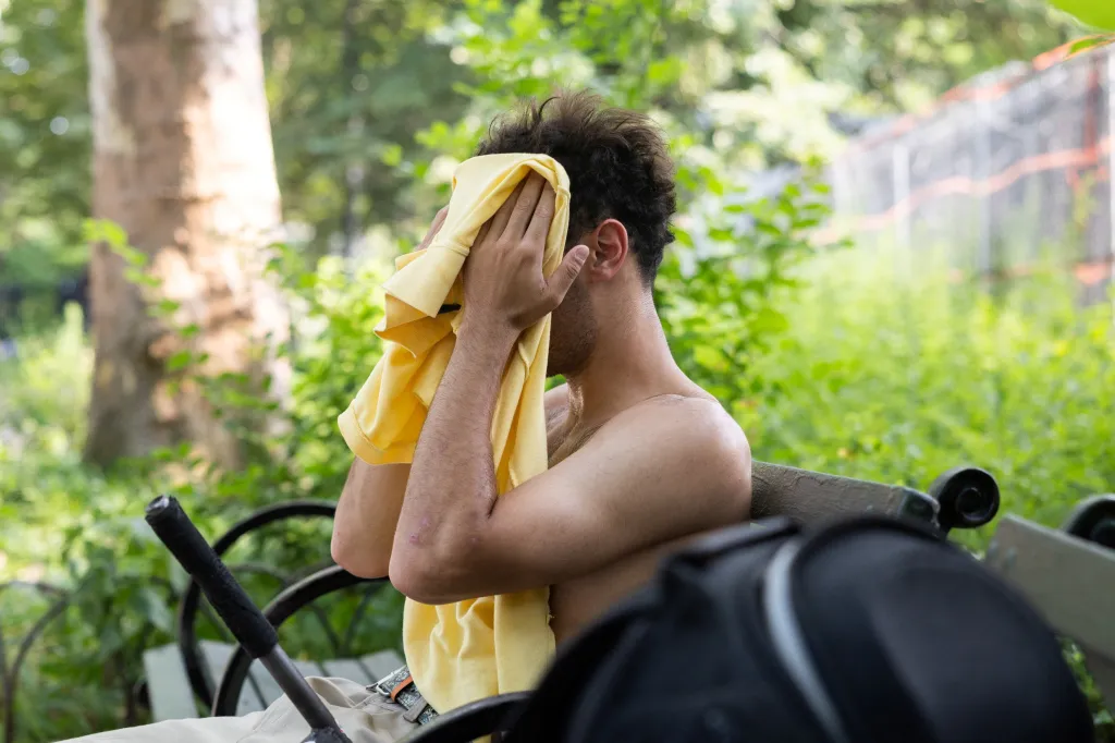 A man sits on a bench, wiping his face with a yellow shirt during a heatwave.