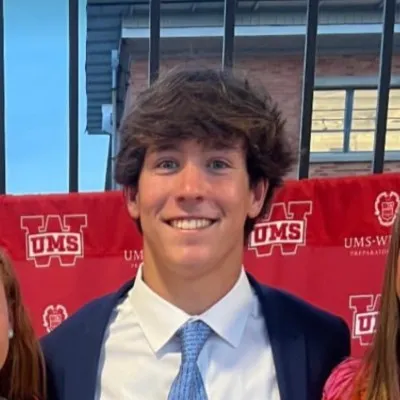 Mark "McNeil" Mostellar smiling, wearing a suit and tie, with a red banner in the background.