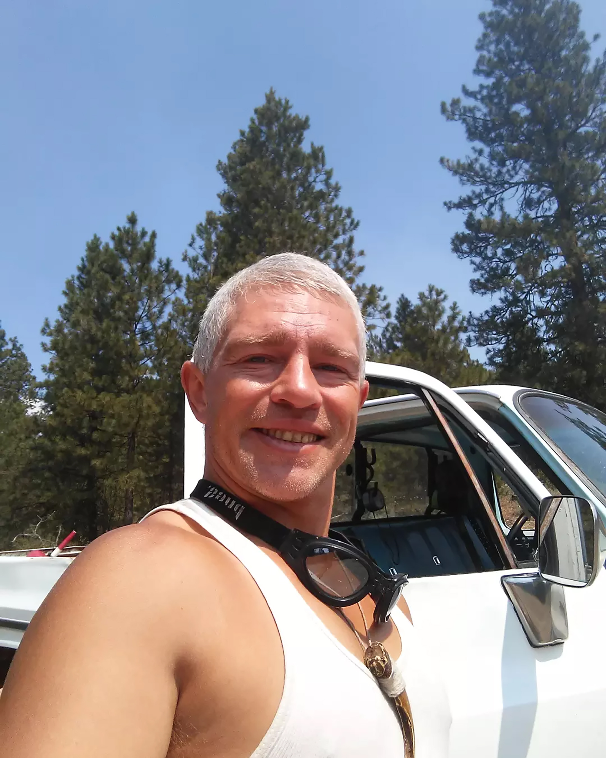 Matt Brown smiling outdoors in a white tank top with goggles around his neck, a white truck and pine trees behind him.
