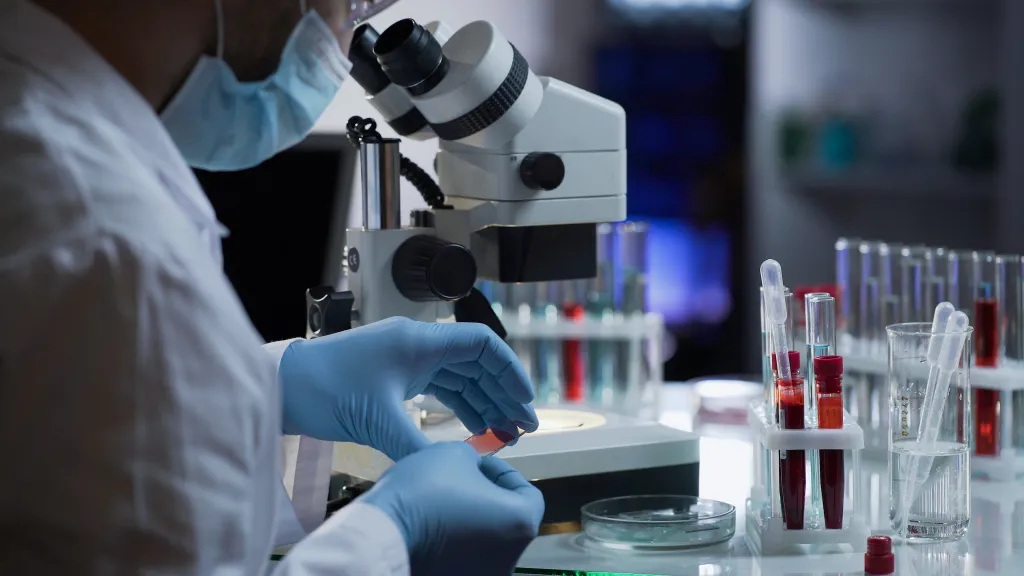 Medical worker in a lab coat and mask analyzing a blood sample under a microscope.