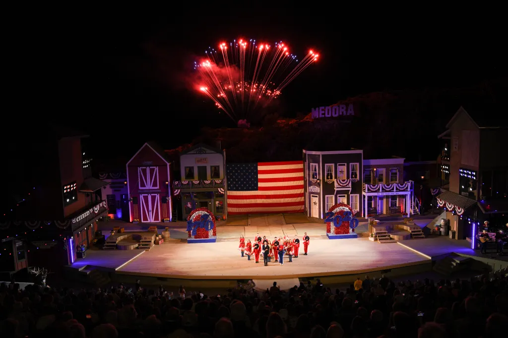 Fireworks light up the night sky over the Medora Musical stage, with performers dressed in red and white on stage.