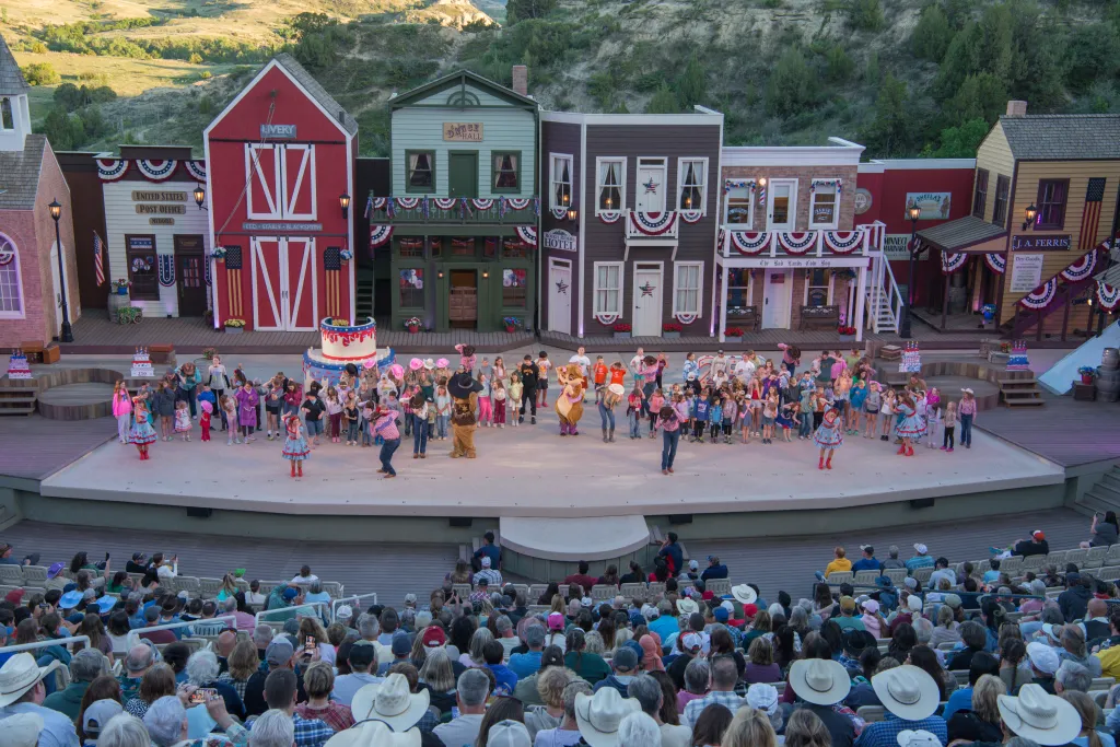 Audience seated in an outdoor amphitheater watching a live performance by a large group of people on stage, set against a backdrop of Old West-style buildings.