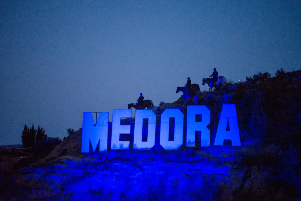 Large illuminated letters spell "Medora" on a hillside, with three silhouetted horse riders visible above them.