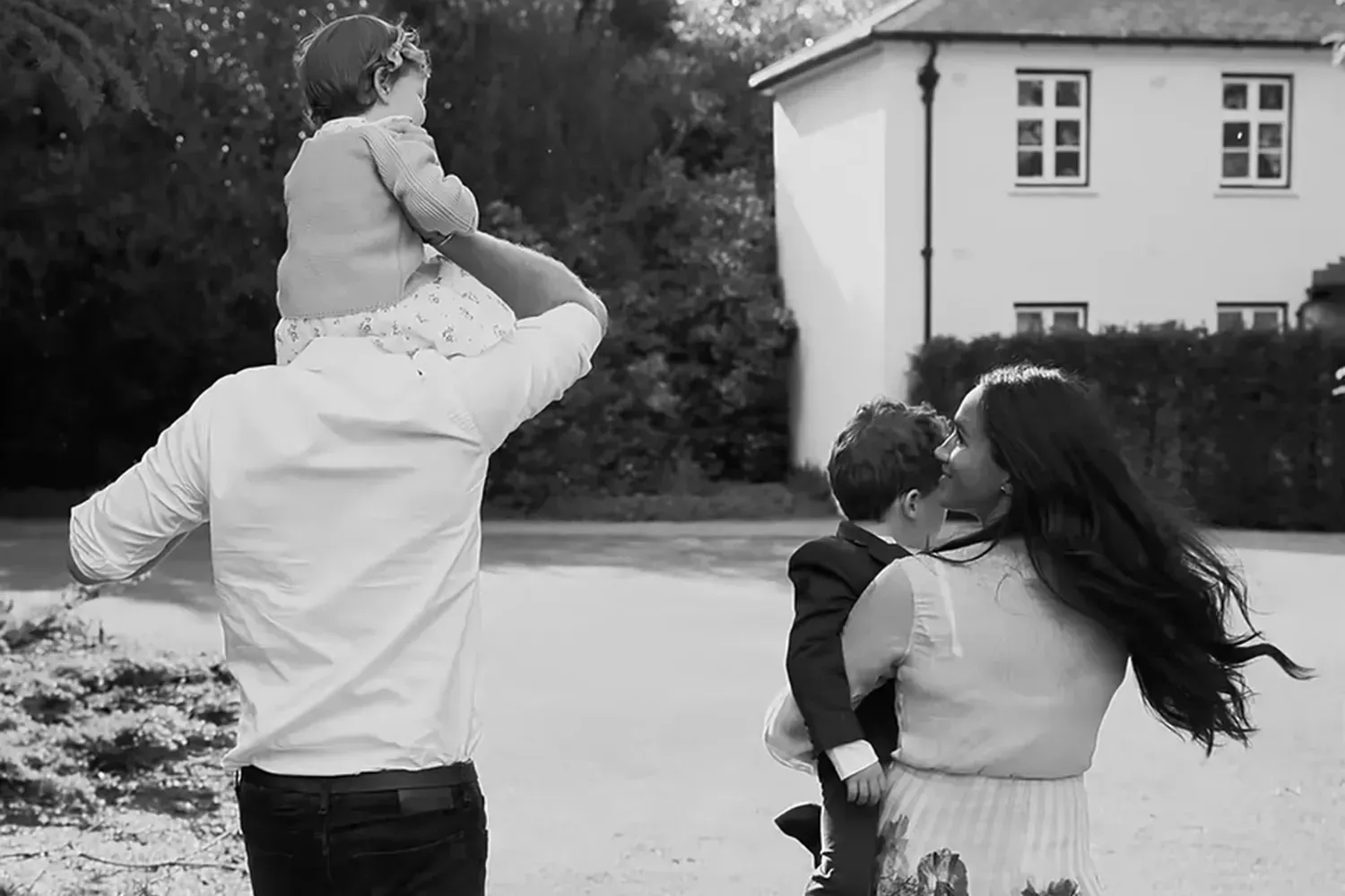 Meghan Markle and Prince Harry with Princess Lilibet and Prince Archie walking away from the camera in black and white.
