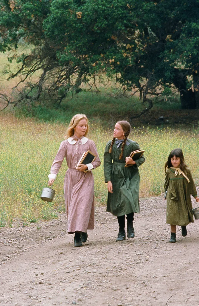 Three girls, Mary, Laura, and Carrie Ingalls, walk down a dirt path.