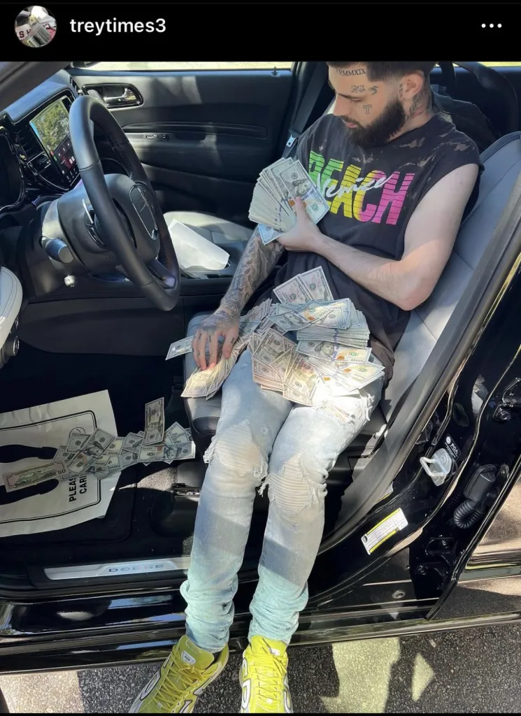 A man with tattoos holding and sitting with stacks of cash in a car.