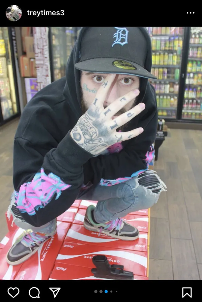 A man with face and hand tattoos, wearing a baseball cap, dark hoodie, and ripped jeans, squats on Coca-Cola boxes with a handgun next to him, covering part of his face with his hand in front of a store's beverage aisle.