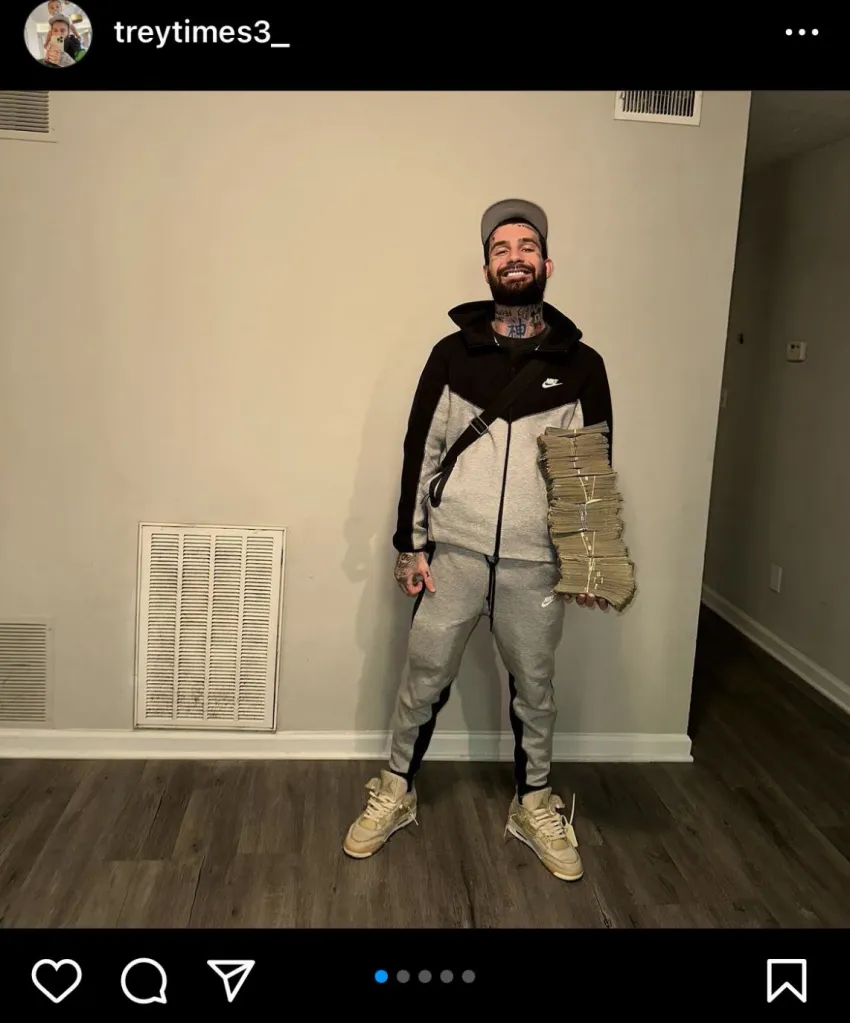 A man in a grey and black tracksuit holds a large stack of money.