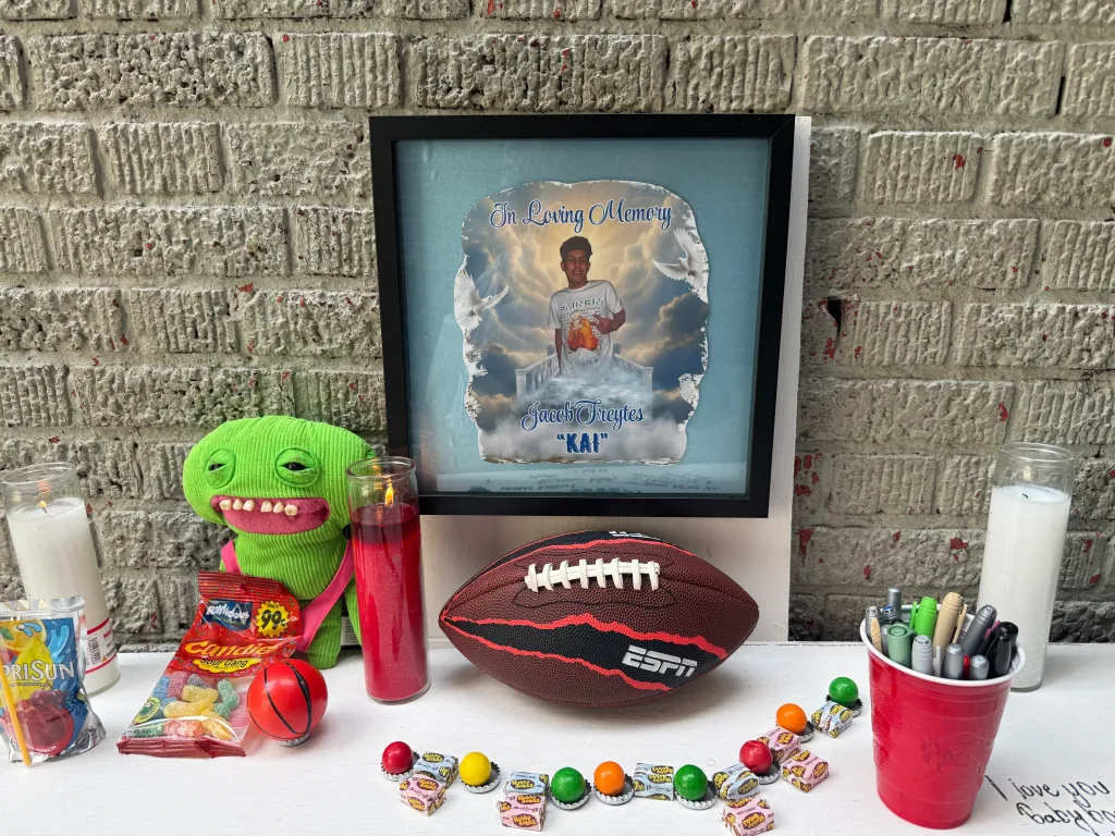 A memorial for Jacob Freytes, featuring a framed photo of him, surrounded by candles, candy, a green plush toy, an ESPN football, and a cup of pens.