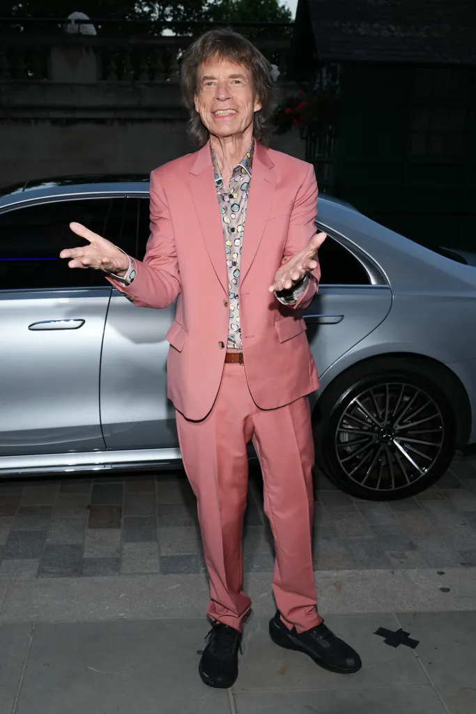 Mick Jagger in a pink suit and patterned shirt, smiling with his hands open.