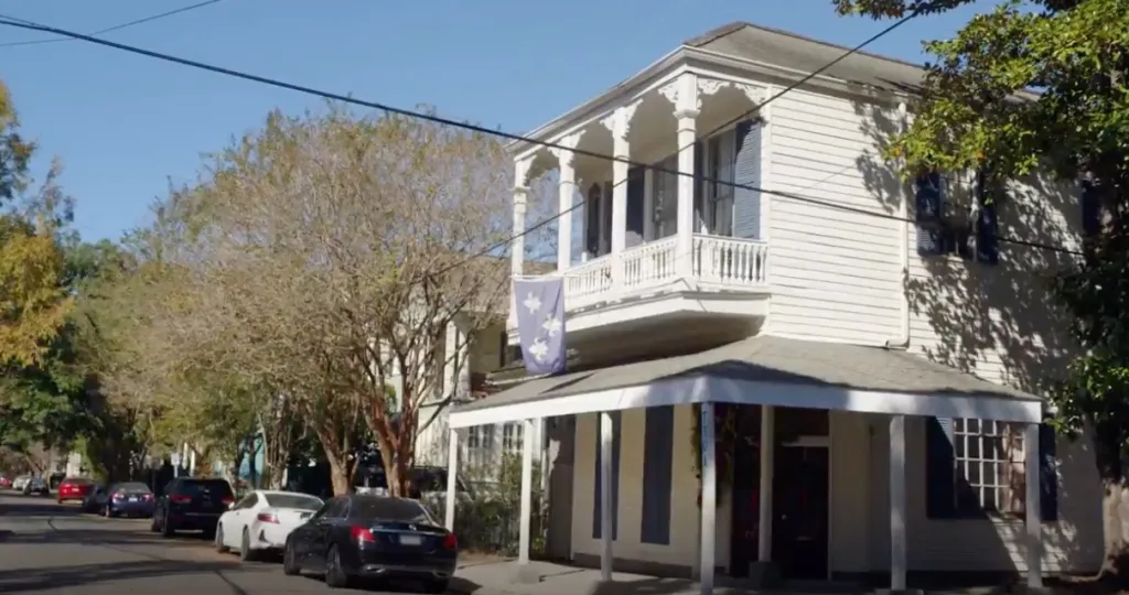 A white two-story house with a balcony and a covered porch, with a blue and white flag hanging from the balcony.
