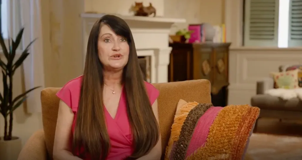 A woman with long dark hair, Debby from 90 Day Fiance, speaks while sitting on a couch.