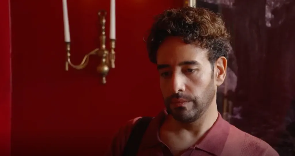 Mido, from 90 Day Fiance, looking down, in a red collared shirt, with a candlestick on a red wall behind him.