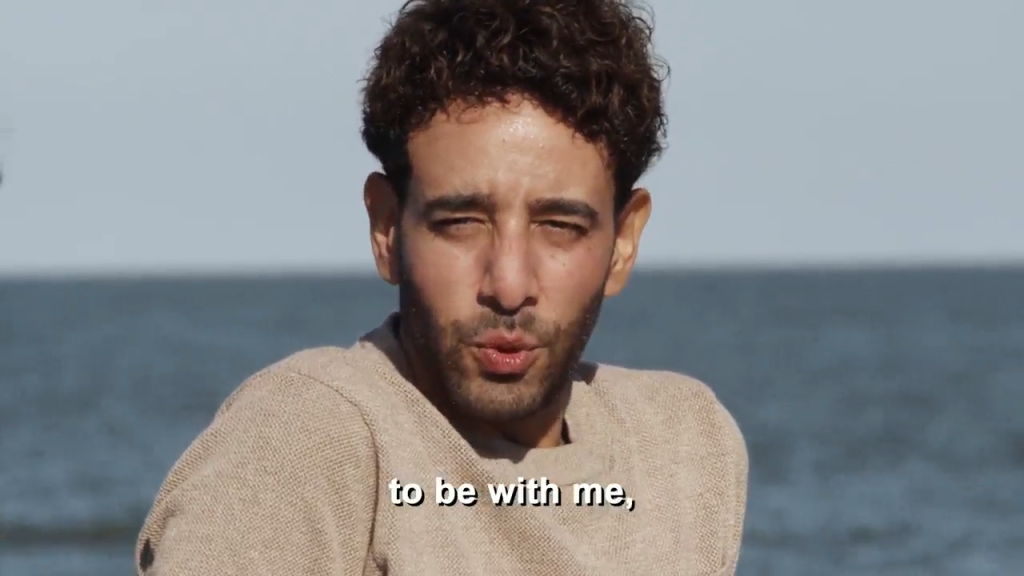 A man with curly hair and a light sweater speaking with the ocean in the background.