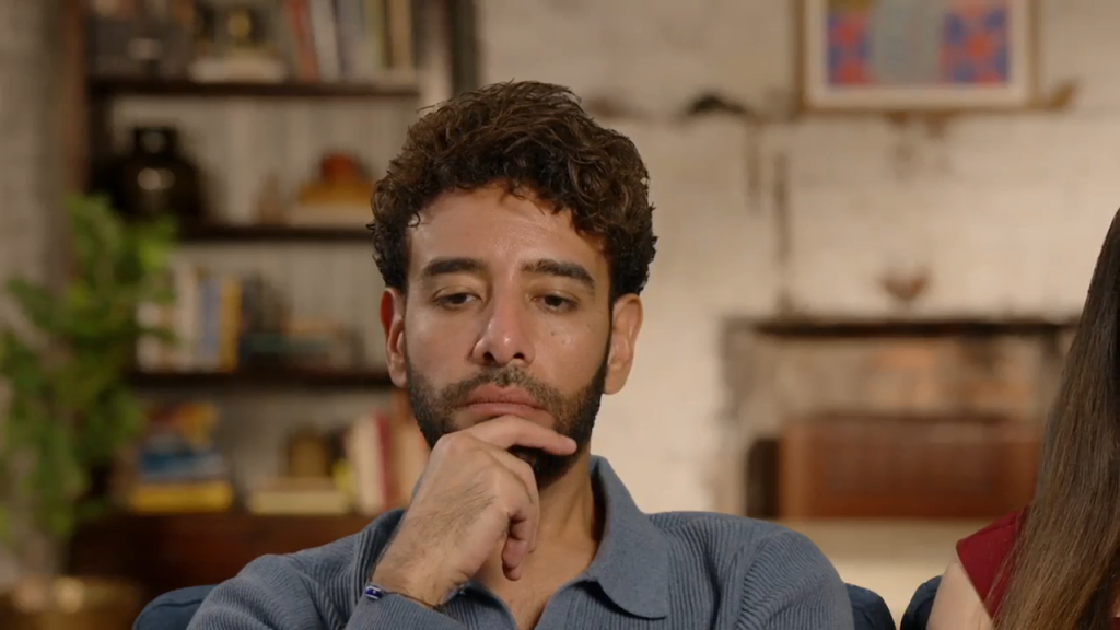 Mido in a light blue ribbed polo, looking down and to the left with his right hand resting on his chin.