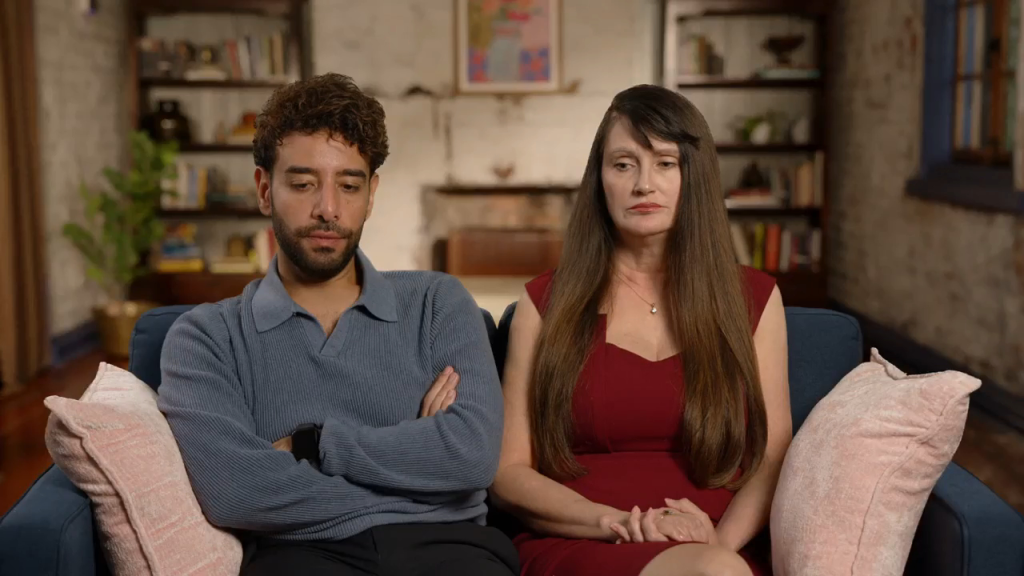 A man with curly hair and a woman with long hair are sitting on a couch, both looking displeased.